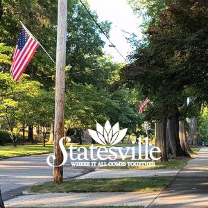 A tree lined street decorated with American flags at dusk. A text overlay reads 'Statesville, Where It All Comes Together'.